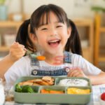 Seorang anak menikmati menu makan siang sehat berisi nasi, ayam, sayuran, telur, dan buah di meja makan dengan suasana ceria. (Foto: dok majalah.sriwijayaterkini.co)